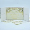 An open, tri-fold **white textured wedding invitation folder** standing on a clean, light surface. The central panel holds the main invitation card, which features elaborate **gold-foil baroque scrollwork** along the top and sides. The text, printed in elegant gold script, announces The side panels of the folder contain pockets for inserts. In the foreground, two coordinating cards—an **RSVP card** and an **Additional Details card**—are laid out, both featuring matching gold-foil borders and modern QR codes. The overall design is regal and sophisticated, with a consistent white and metallic gold color palette.