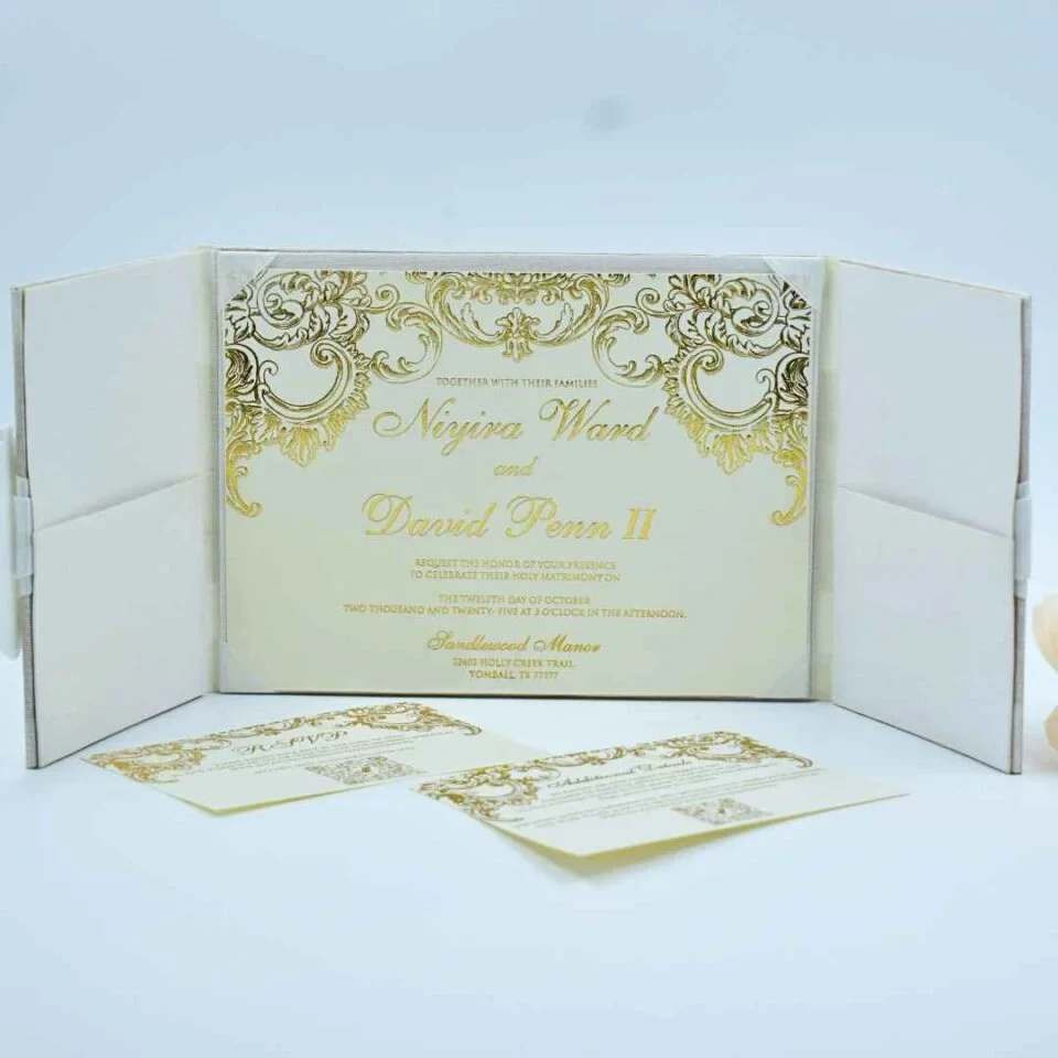 An open, tri-fold **white textured wedding invitation folder** standing on a clean, light surface. The central panel holds the main invitation card, which features elaborate **gold-foil baroque scrollwork** along the top and sides. The text, printed in elegant gold script, announces The side panels of the folder contain pockets for inserts. In the foreground, two coordinating cards—an **RSVP card** and an **Additional Details card**—are laid out, both featuring matching gold-foil borders and modern QR codes. The overall design is regal and sophisticated, with a consistent white and metallic gold color palette.