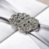 A detailed, macro close-up of a shimmering **rhinestone crystal brooch** centered on a luxurious wedding invitation. The brooch features an intricate, scalloped filigree design packed with small, sparkling clear stones. It is fastened over a wide, **silver-grey satin ribbon** that has a soft, metallic sheen. The ribbon sits atop a **white silk-textured box**, with the fine horizontal weave of the fabric visible in the shallow depth of field. The lighting is bright and focused, highlighting the reflective facets of the crystals and the smooth texture of the ribbon.