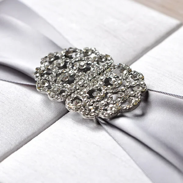 A detailed, macro close-up of a shimmering **rhinestone crystal brooch** centered on a luxurious wedding invitation. The brooch features an intricate, scalloped filigree design packed with small, sparkling clear stones. It is fastened over a wide, **silver-grey satin ribbon** that has a soft, metallic sheen. The ribbon sits atop a **white silk-textured box**, with the fine horizontal weave of the fabric visible in the shallow depth of field. The lighting is bright and focused, highlighting the reflective facets of the crystals and the smooth texture of the ribbon.