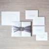 A top-down, flat-lay view of a complete white and silver wedding invitation suite arranged on a light wood-grain surface. The centerpiece is a square, white silk-textured invitation box cinched with a wide, silver-grey satin ribbon and a sparkling rhinestone filigree brooch. Surrounding the box are several coordinating stationery pieces printed in elegant black script.