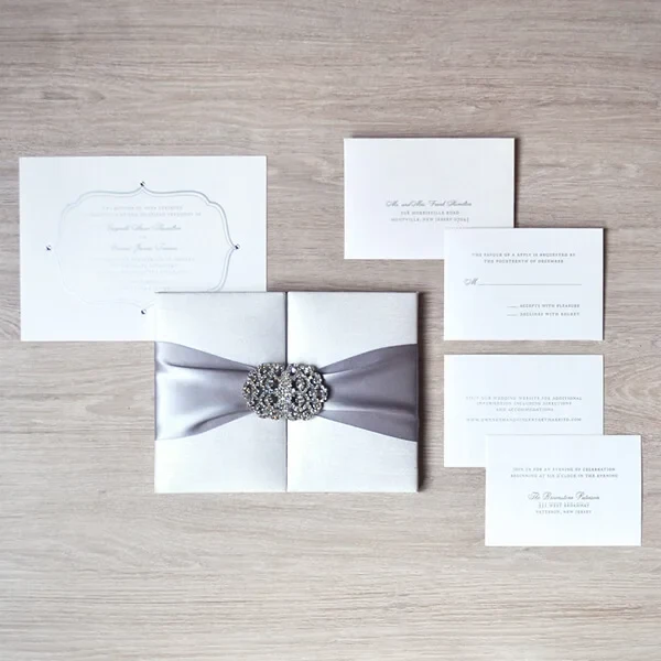 A top-down, flat-lay view of a complete white and silver wedding invitation suite arranged on a light wood-grain surface. The centerpiece is a square, white silk-textured invitation box cinched with a wide, silver-grey satin ribbon and a sparkling rhinestone filigree brooch. Surrounding the box are several coordinating stationery pieces printed in elegant black script.