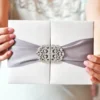 A close-up shot of a person with pink manicured nails holding a luxurious, white silk-textured wedding invitation box. The box is secured with a wide, dusty lavender or silver-grey satin ribbon that wraps horizontally around the center. At the midpoint, the ribbon is cinched by a large, ornate rhinestone crystal brooch with a filigree design, adding a sophisticated sparkle. The person holding the invitation wears a light-colored, embellished garment, softly blurred in the background.