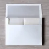 A top-down view of an open **white mailing envelope** resting on a light wood-grain surface. Inside the envelope, the **white silk wedding invitation box** is tucked securely, with its shimmering, textured fabric visible. The envelope features a **silver-grey inner liner** on the open flap, coordinating with the silver accents of the invitation suite. The presentation is clean and organized, showing how the bulky invitation box fits perfectly within its custom mailing packaging.