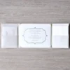 A top-down view of the **white silk wedding invitation folder** fully opened on a light wood-grain surface, displaying its tri-fold design. The central panel holds the **main invitation card** securely in place with four silk corner pockets. The card features a classic silver-embossed decorative frame and lists the names of the couple. On either side of the center, the left and right panels feature **built-in horizontal pockets** that hold coordinating insert cards, such as the RSVP and detail cards, neatly tucked inside. The entire suite maintains a clean, monochromatic white and silver aesthetic.