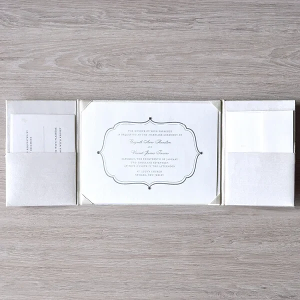 A top-down view of the **white silk wedding invitation folder** fully opened on a light wood-grain surface, displaying its tri-fold design. The central panel holds the **main invitation card** securely in place with four silk corner pockets. The card features a classic silver-embossed decorative frame and lists the names of the couple. On either side of the center, the left and right panels feature **built-in horizontal pockets** that hold coordinating insert cards, such as the RSVP and detail cards, neatly tucked inside. The entire suite maintains a clean, monochromatic white and silver aesthetic.