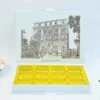 An open, white luxury wedding invittaion box featuring a detailed, gold-etched illustration of a grand historic building on the inner lid. Beneath the building, the name Grand Hotel Excelsior Vittoria is printed in an elegant gold script. The base of the box contains a bright gold-coclate tray with twelve individual rectangular compartments, designed to hold chocolates or small treats. The box is set against a clean, light blue background with a hint of a peach flower on the far left.