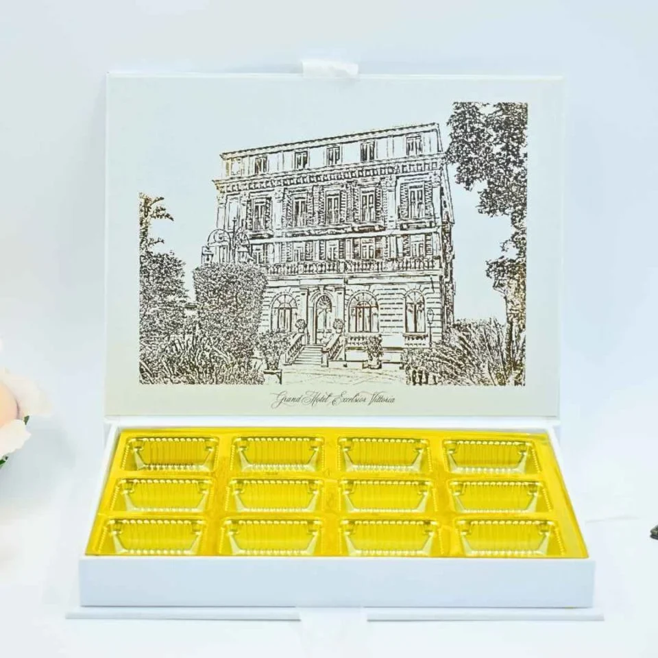 An open, white luxury wedding invittaion box featuring a detailed, gold-etched illustration of a grand historic building on the inner lid. Beneath the building, the name Grand Hotel Excelsior Vittoria is printed in an elegant gold script. The base of the box contains a bright gold-coclate tray with twelve individual rectangular compartments, designed to hold chocolates or small treats. The box is set against a clean, light blue background with a hint of a peach flower on the far left.
