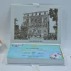 An open, white luxury wedding invittaion box featuring a detailed, gold-etched illustration of a grand historic building on the inner lid. Beneath the building, the name Grand Hotel Excelsior Vittoria is printed in an elegant gold script. The base of the box contains a bright gold-coclate tray with twelve individual rectangular compartments, designed to hold chocolates or small treats. The box is set against a clean, light blue background with a hint of a peach flower on the far left.