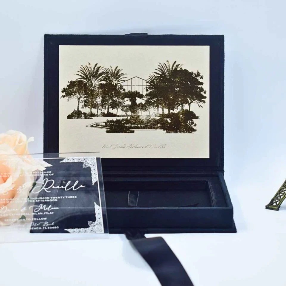An open, velvet wedding invitation box displayed against a light blue background. The inner lid features a sepia-toned etched illustration of a lush botanical garden with palm trees and a glass conservatory. To the left, a clear acrylic invitation with white script lettering leans against a soft peach rose. The box features a deep recessed compartment in the base, and a matching navy blue satin ribbon is visible at the bottom. A small bronze-colored stands to the right, completing the elegant travel-themed presentation.