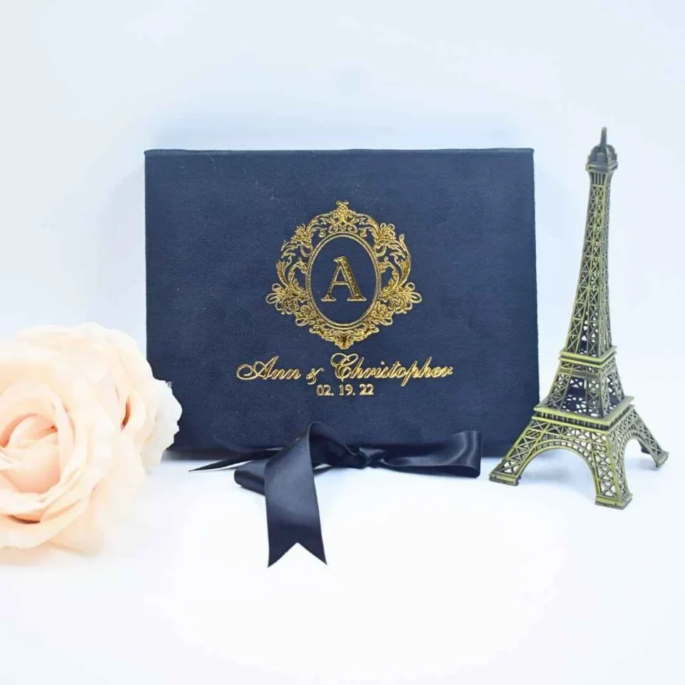 A front-facing view of a rectangular, velvet wedding invitation box set against a light blue background. The lid is detailed with a shimmering gold foil crest containing the letter in the center. Below the crest, the name and the date are printed in an elegant gold script. The box is styled with: A matching satin ribbon tied in a bow at the bottom. A small, dark bronze Eiffel Tower figurine standing to the right. A soft, cream-colored rose partially visible on the left side of the frame.