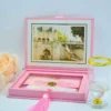 A high-angle view of an open velvet wedding invitation box displayed against a light blue background. The luxurious set includes:Inside Lid:A stunning gold-foiled illustration of a grand, historic building with palm trees and a detailed staircase.The Base A clear acrylic invitation card featuring white and gold script text, accented by a thick burgundy silk tassel.Stationery: Coordinating white cards for "Details" and "Accommodations" with gold-foiled floral scrollwork are scattered around the box.Decorative Elements:** A small, dark bronze Eiffel Tower figurine stands to the right, and a soft peach-colored rose is visible on the left. A long satin ribbon weaves through the arrangement, completing the elegant and regal aesthetic.