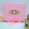 A front-facing view of a closed, rectangular dark emerald green velvet wedding invitation box set against a clean white background. The sophisticated design includes: * A large, central gold foil monogram on the lid featuring the initials in an elegant script, topped with a detailed royal crown. An intricate gold foil border consisting of ornate floral scrollwork that frames the edges of the box. Decorative accents in the background, including a soft peach-colored rose on the left and a small, dark bronze Eiffel Tower figurine** on the right. The contrast between the deep green velvet and the shimmering gold foil creates a regal and luxurious aesthetic.