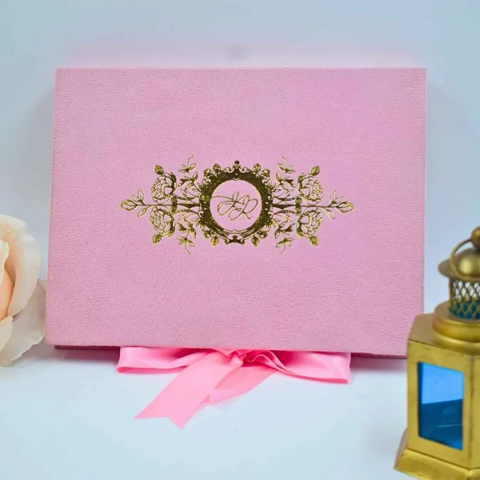 A front-facing view of a closed, rectangular dark emerald green velvet wedding invitation box set against a clean white background. The sophisticated design includes: * A large, central gold foil monogram on the lid featuring the initials in an elegant script, topped with a detailed royal crown. An intricate gold foil border consisting of ornate floral scrollwork that frames the edges of the box. Decorative accents in the background, including a soft peach-colored rose on the left and a small, dark bronze Eiffel Tower figurine** on the right. The contrast between the deep green velvet and the shimmering gold foil creates a regal and luxurious aesthetic.