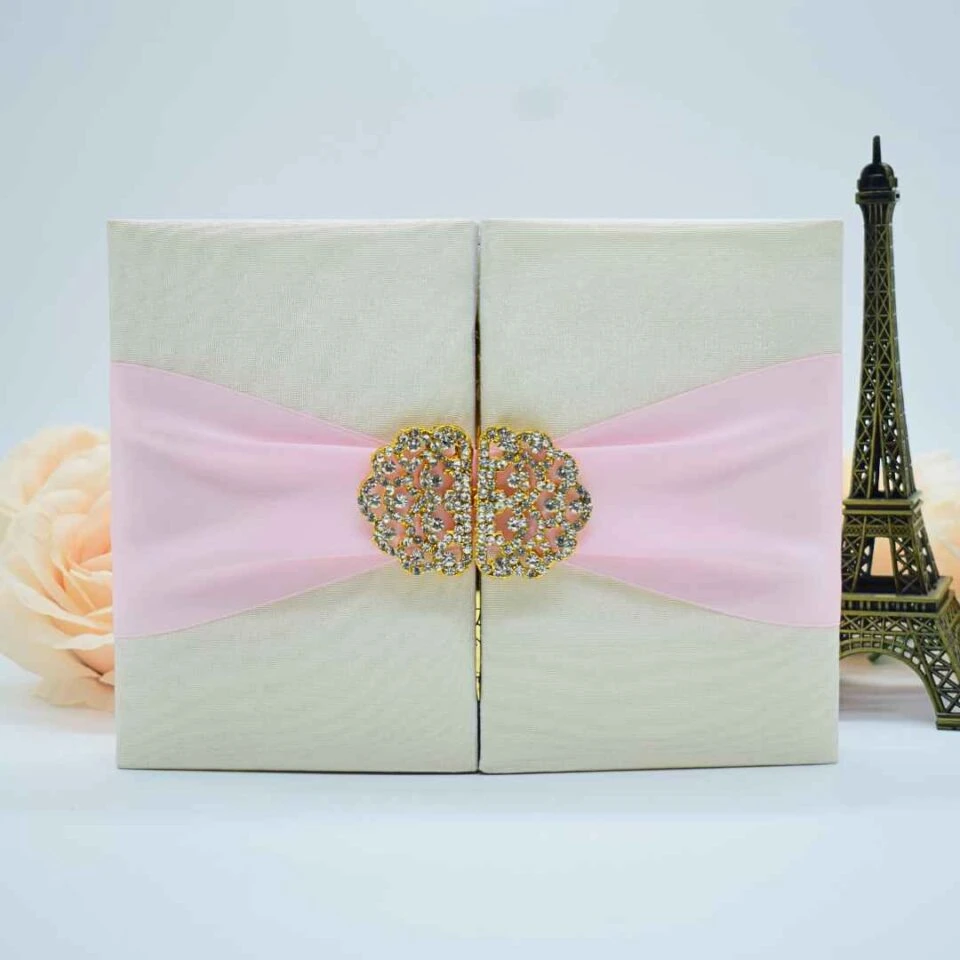 A front-facing shot of a **closed cream-colored gatefold invitation box** set against a minimalist white background. The box features: * A **wide pink silk sash** wrapped horizontally around the center. * A large, two-piece **gold filigree brooch** encrusted with sparkling clear crystals, acting as a decorative clasp where the gatefold doors meet. * The box has a subtle, fine-grained fabric texture. * Decorative accents in the background include a small **bronze Eiffel Tower figurine** on the right and a soft peach-colored rose on the left. * The overall aesthetic is elegant and romantic, with a focus on the shimmering brooch and soft color palette.