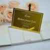 An open, **luxurious cream-colored gatefold invitation box** displaying its interior contents against a bright white background. The suite includes: * **Interior Panels:** Both inner "doors" of the gatefold feature horizontal pockets holding cream-colored RSVP or detail cards with elegant gold accents. * **The Centerpiece:** A **gold mirrored acrylic invitation card** sits in the main compartment, etched with white calligraphy for "Maria Goretti Numfor and Oscar C. Ambe." The card is embellished with small, decorative gold studs. * **Exterior Detail:** The top of the box features a **wide pink silk sash** and a large, intricate **gold filigree brooch** encrusted with crystals. * **Decor:** The arrangement is accented by a small **bronze Eiffel Tower figurine** and a cluster of soft peach and white roses in the background.