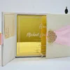 An open, **luxurious cream-colored gatefold invitation box** displaying its interior contents against a bright white background. The suite includes: * **Interior Panels:** Both inner "doors" of the gatefold feature horizontal pockets holding cream-colored RSVP or detail cards with elegant gold accents. * **The Centerpiece:** A **gold mirrored acrylic invitation card** sits in the main compartment, etched with white calligraphy for "Maria Goretti Numfor and Oscar C. Ambe." The card is embellished with small, decorative gold studs. * **Exterior Detail:** The top of the box features a **wide pink silk sash** and a large, intricate **gold filigree brooch** encrusted with crystals. * **Decor:** The arrangement is accented by a small **bronze Eiffel Tower figurine** and a cluster of soft peach and white roses in the background.