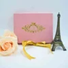 A rectangular, dusty rose velvet invitation box stands upright against a white background. The center of the box is embellished with an intricate gold foil crest featuring a crown and ornate scrollwork. In front of the box lies a loosely tied gold satin ribbon. To the left, a soft peach-colored rose is partially visible, and to the right stands a small, dark bronze Eiffel Tower figurine.