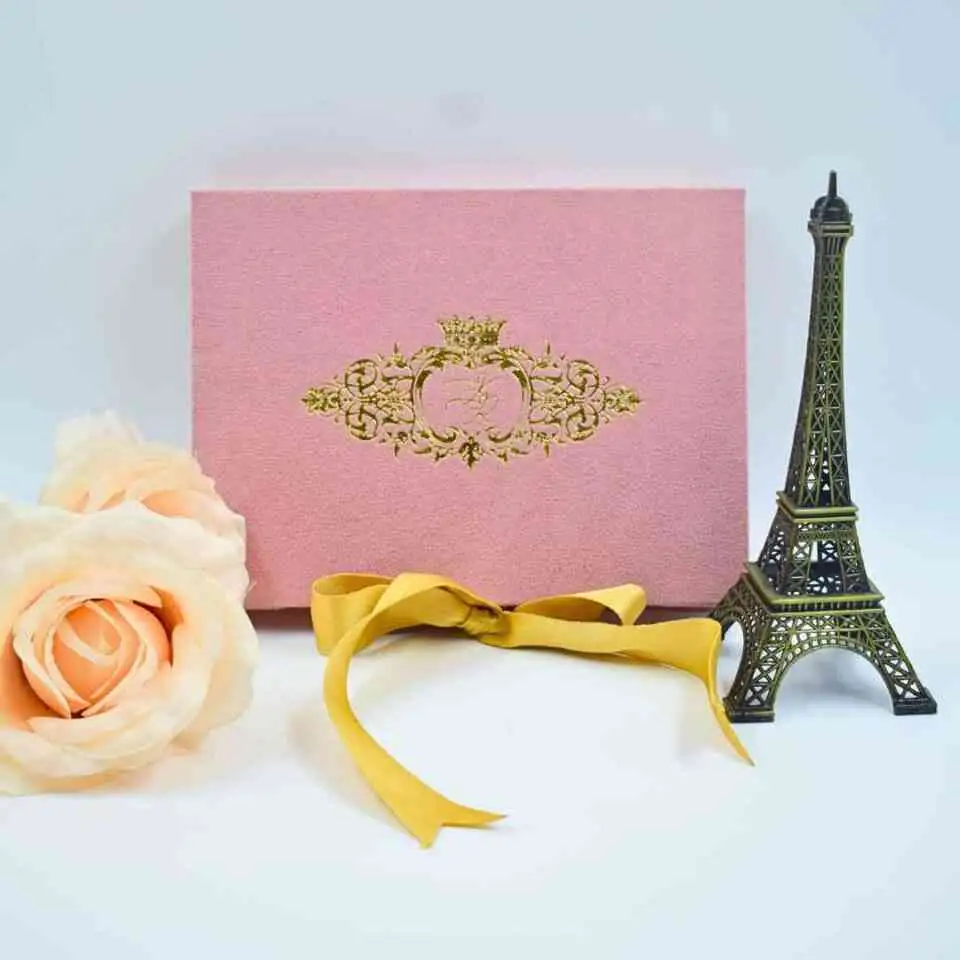 A rectangular, dusty rose velvet invitation box stands upright against a white background. The center of the box is embellished with an intricate gold foil crest featuring a crown and ornate scrollwork. In front of the box lies a loosely tied gold satin ribbon. To the left, a soft peach-colored rose is partially visible, and to the right stands a small, dark bronze Eiffel Tower figurine.