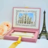 A square, dusty rose velvet invitation box is shown open on a white surface. The interior of the lid features a matching velvet pocket holding an ivory card with a gold-foil crown and monogram crest. In the base of the box, a formal invitation card for a "Sweet 16" celebration for Arlyn Estrella Bac is displayed with gold-stamped scrollwork at the corners. To the left, a soft peach rose is visible, and to the right stands a small bronze Eiffel Tower figurine.