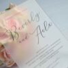 An elegant, frosted acrylic wedding invitation is displayed at an angle, resting against a bouquet of soft peach and cream roses. The frosted material of the invitation creates a soft, semi-transparent effect, allowing the delicate petals of the roses behind it to peek through.