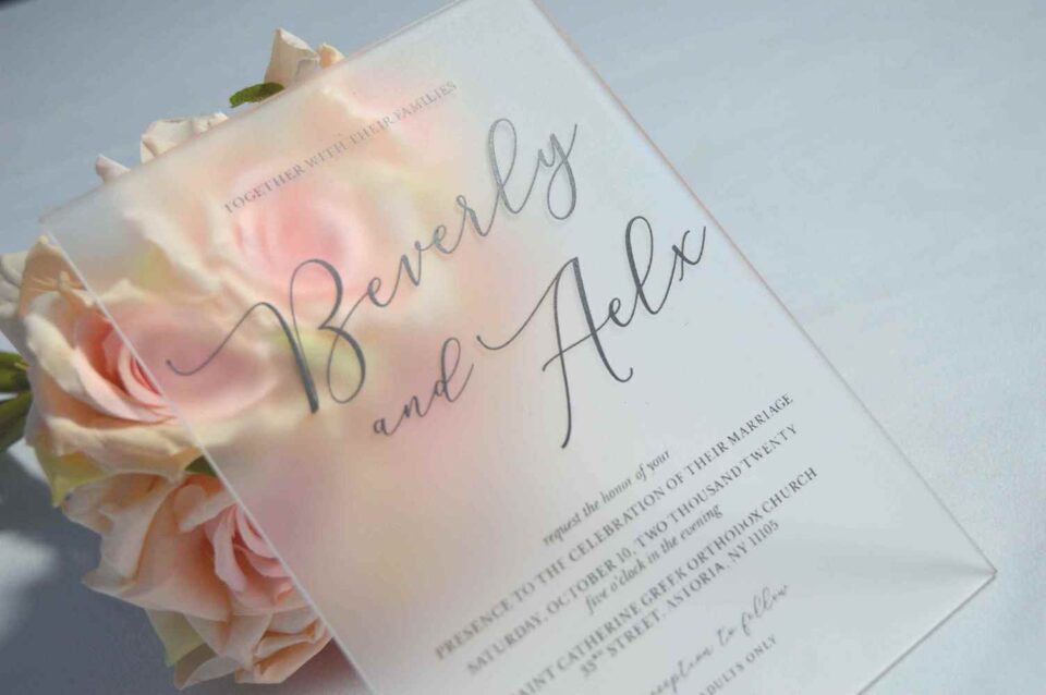 An elegant, frosted acrylic wedding invitation is displayed at an angle, resting against a bouquet of soft peach and cream roses. The frosted material of the invitation creates a soft, semi-transparent effect, allowing the delicate petals of the roses behind it to peek through.