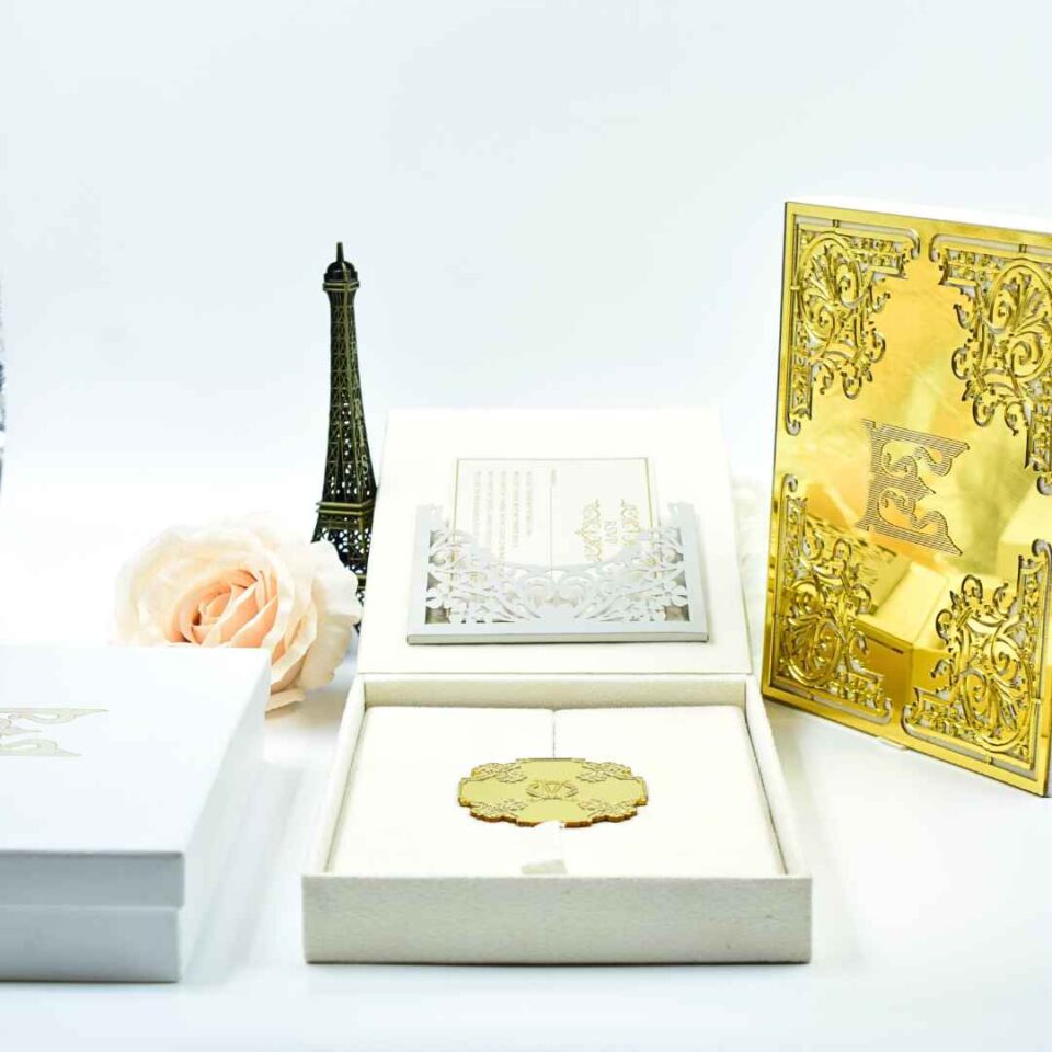 A luxury ivory-colored wedding invitation suite featuring a laser-cut gold acrylic folio, an open invitation box with an ornate gold mirror acrylic monogram centerpiece, and elegant floral-patterned card holders, displayed with a silk rose.