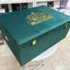 A left-side view of a luxury emerald green velvet bridal lehenga box, featuring a crown-style gold mirror acrylic laser-cut monogram on the lid and two gold metal locks on the front, set against a retail display background.