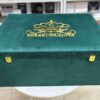 A high-angle view of a luxury emerald green velvet bridal lehenga box, featuring a crown-style gold mirror acrylic laser-cut monogram on the lid and two gold metal locks on the front, set against a retail display background.