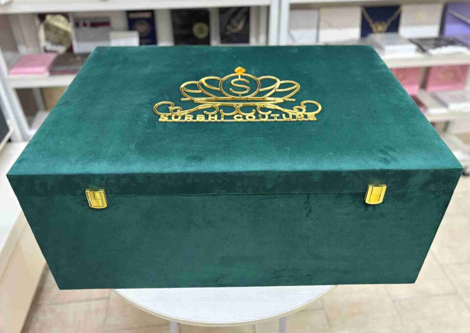 A high-angle view of a luxury emerald green velvet bridal lehenga box, featuring a crown-style gold mirror acrylic laser-cut monogram on the lid and two gold metal locks on the front, set against a retail display background.
