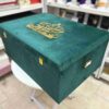 A right-side view of a luxury emerald green velvet bridal lehenga box, featuring a crown-style gold mirror acrylic laser-cut monogram on the lid and two gold metal locks on the front, set against a retail display background.