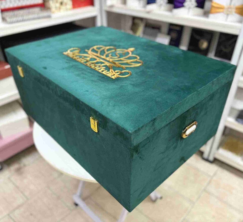 A right-side view of a luxury emerald green velvet bridal lehenga box, featuring a crown-style gold mirror acrylic laser-cut monogram on the lid and two gold metal locks on the front, set against a retail display background.