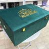 A left-side view of a luxury emerald green velvet bridal lehenga box, featuring a crown-style gold mirror acrylic laser-cut monogram on the lid and two gold metal locks on the front, set against a retail display background.