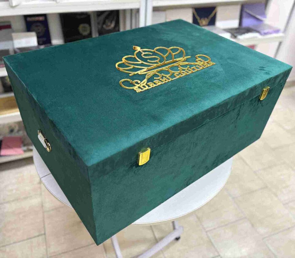 A left-side view of a luxury emerald green velvet bridal lehenga box, featuring a crown-style gold mirror acrylic laser-cut monogram on the lid and two gold metal locks on the front, set against a retail display background.