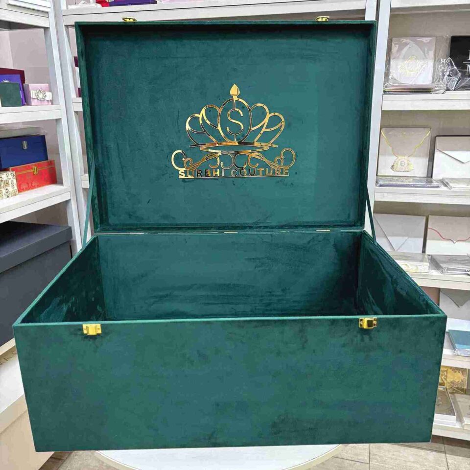 A high-angle open view of a luxury emerald green velvet bridal lehenga box, featuring a crown-style gold mirror acrylic laser-cut monogram on the lid and two gold metal locks on the front, set against a retail display background.