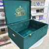 A high-angle open view of a luxury emerald green velvet bridal lehenga box, featuring a crown-style gold mirror acrylic laser-cut monogram on the lid and two gold metal locks on the front, set against a retail display background.