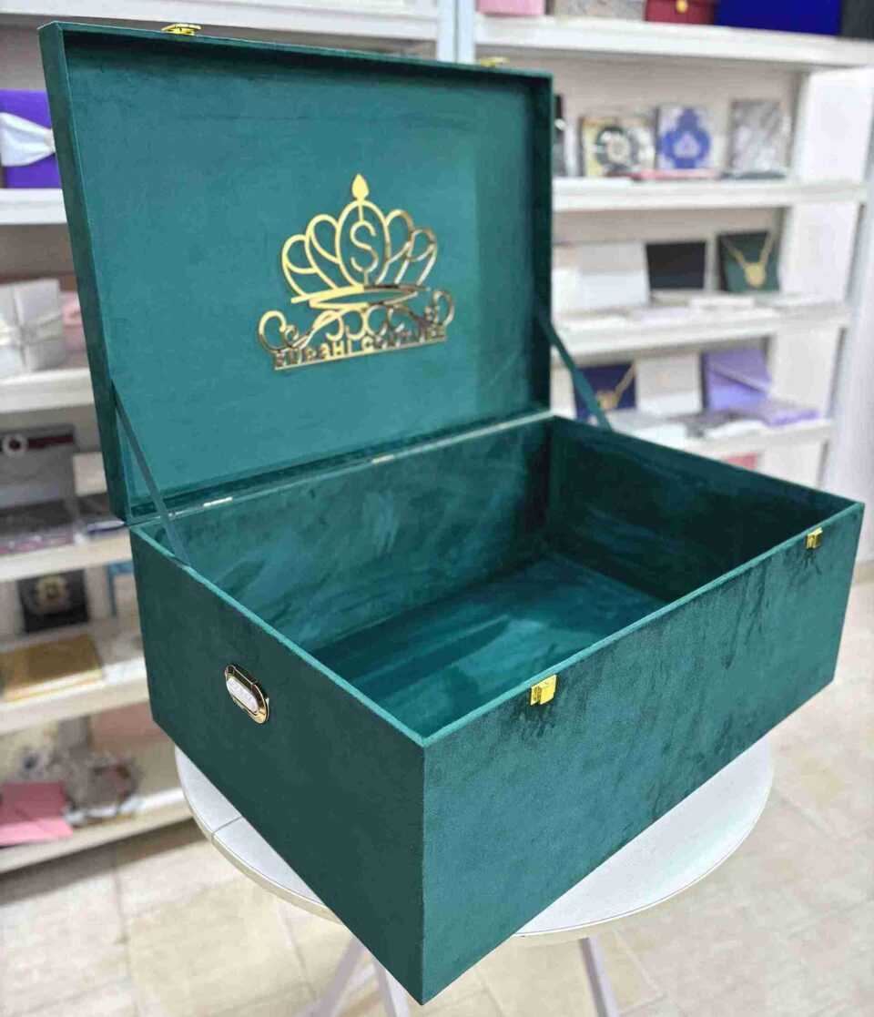 A high-angle open view of a luxury emerald green velvet bridal lehenga box, featuring a crown-style gold mirror acrylic laser-cut monogram on the lid and two gold metal locks on the front, set against a retail display background.
