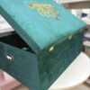 A high-angle semi-open back view of a luxury emerald green velvet bridal lehenga box, featuring a crown-style gold mirror acrylic laser-cut monogram on the lid and two gold metal locks on the front, set against a retail display background.