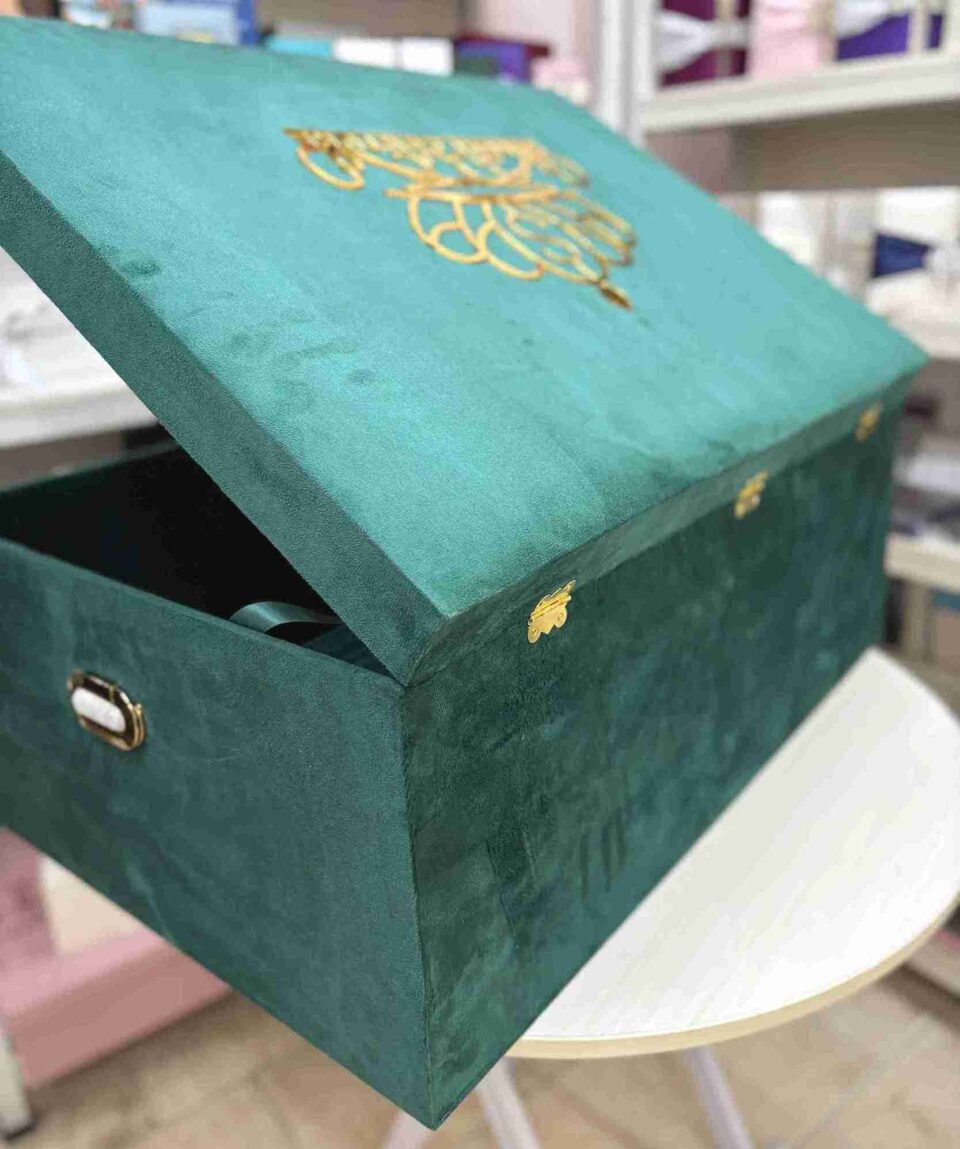A high-angle semi-open back view of a luxury emerald green velvet bridal lehenga box, featuring a crown-style gold mirror acrylic laser-cut monogram on the lid and two gold metal locks on the front, set against a retail display background.
