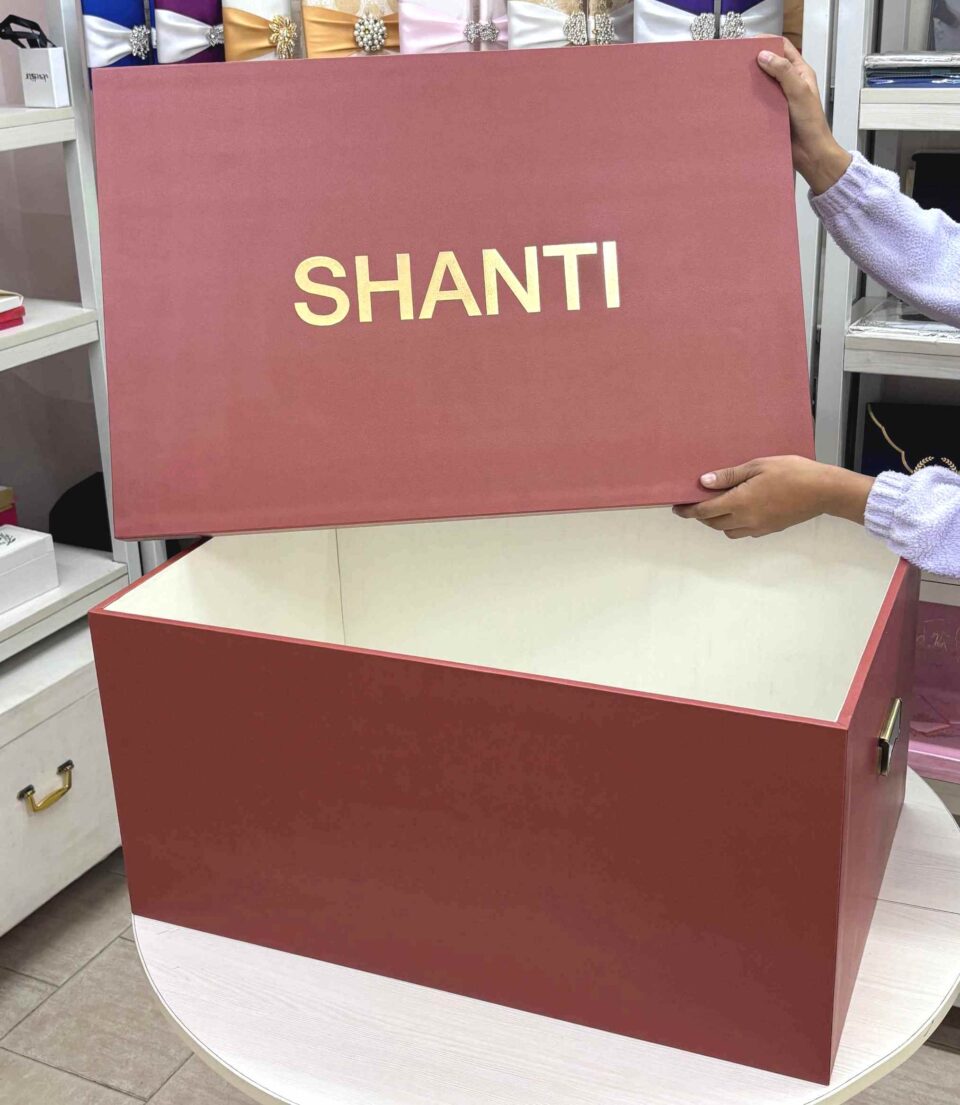 A high-end bridal lehenga trunk box made of a thick orange MDF frame with a smooth matte finish, featuring a custom gold foil-pressed logo on the lid and a sturdy antique brass handle on the side for professional fashion packaging.