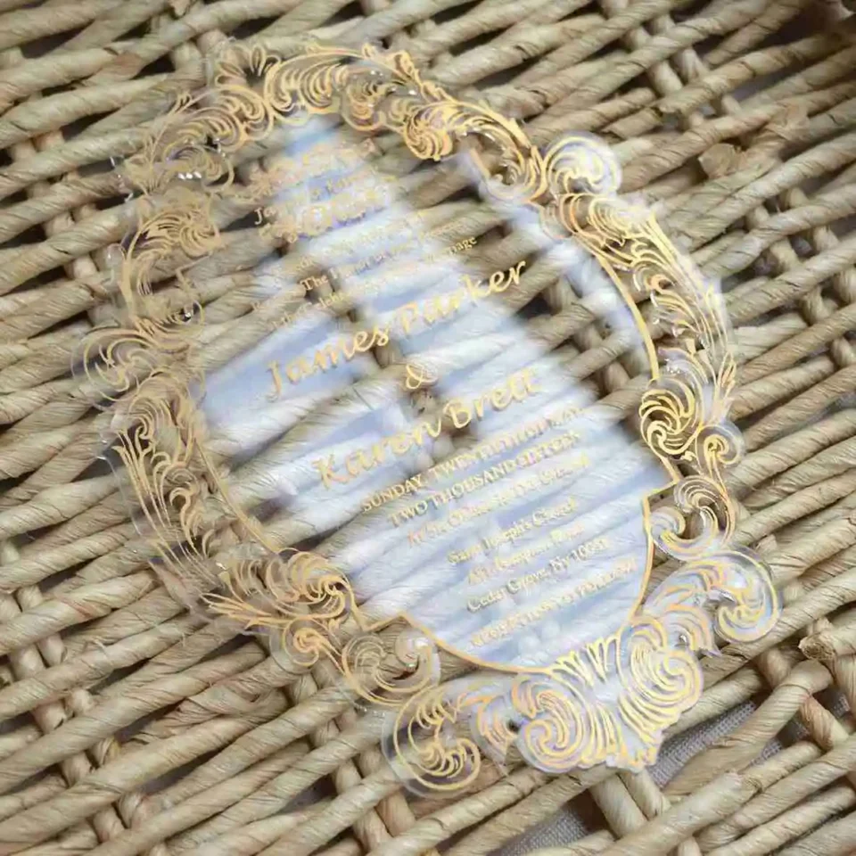 Clear acrylic wedding invitation with gold baroque frame, laid on a woven rattan surface
