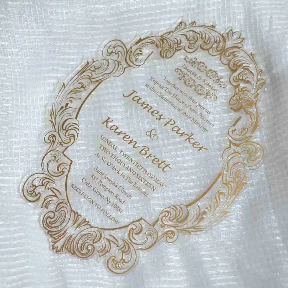 Close-up of clear acrylic invitation with gold engraved baroque scrollwork border on white linen