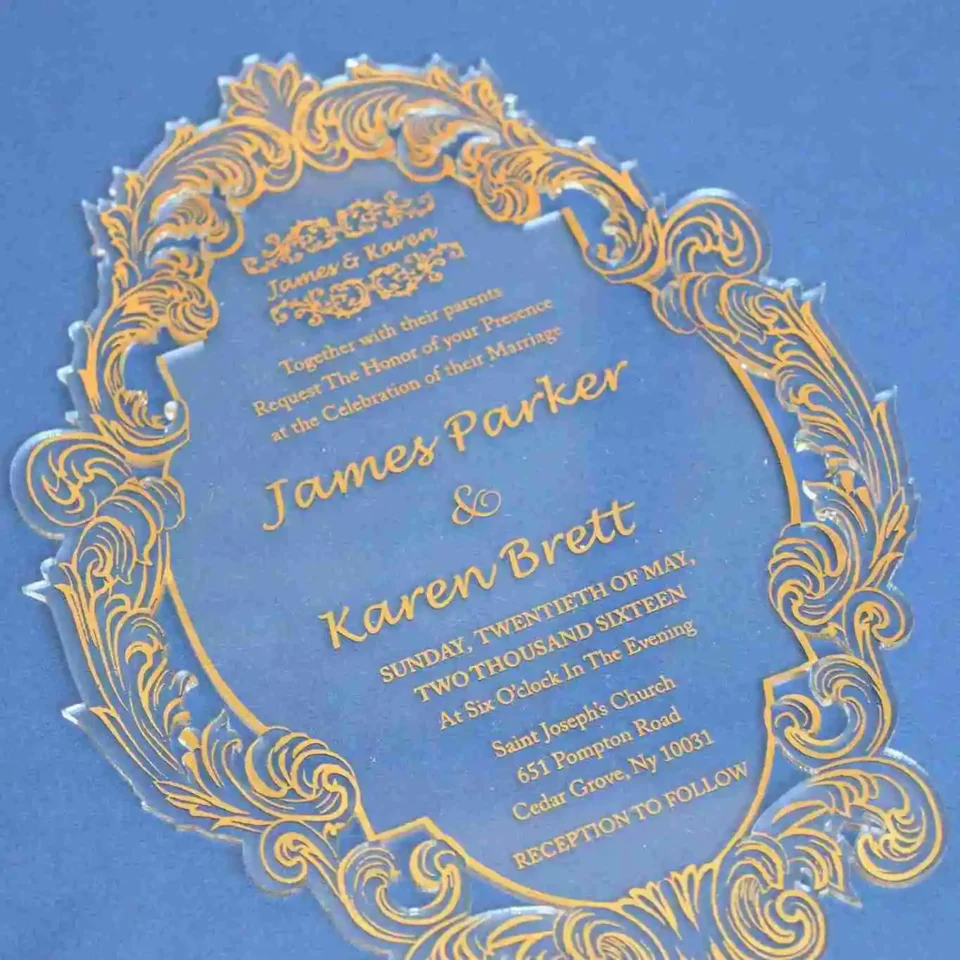 Clear acrylic wedding invitation with gold ornate frame, angled view against a blue backdrop