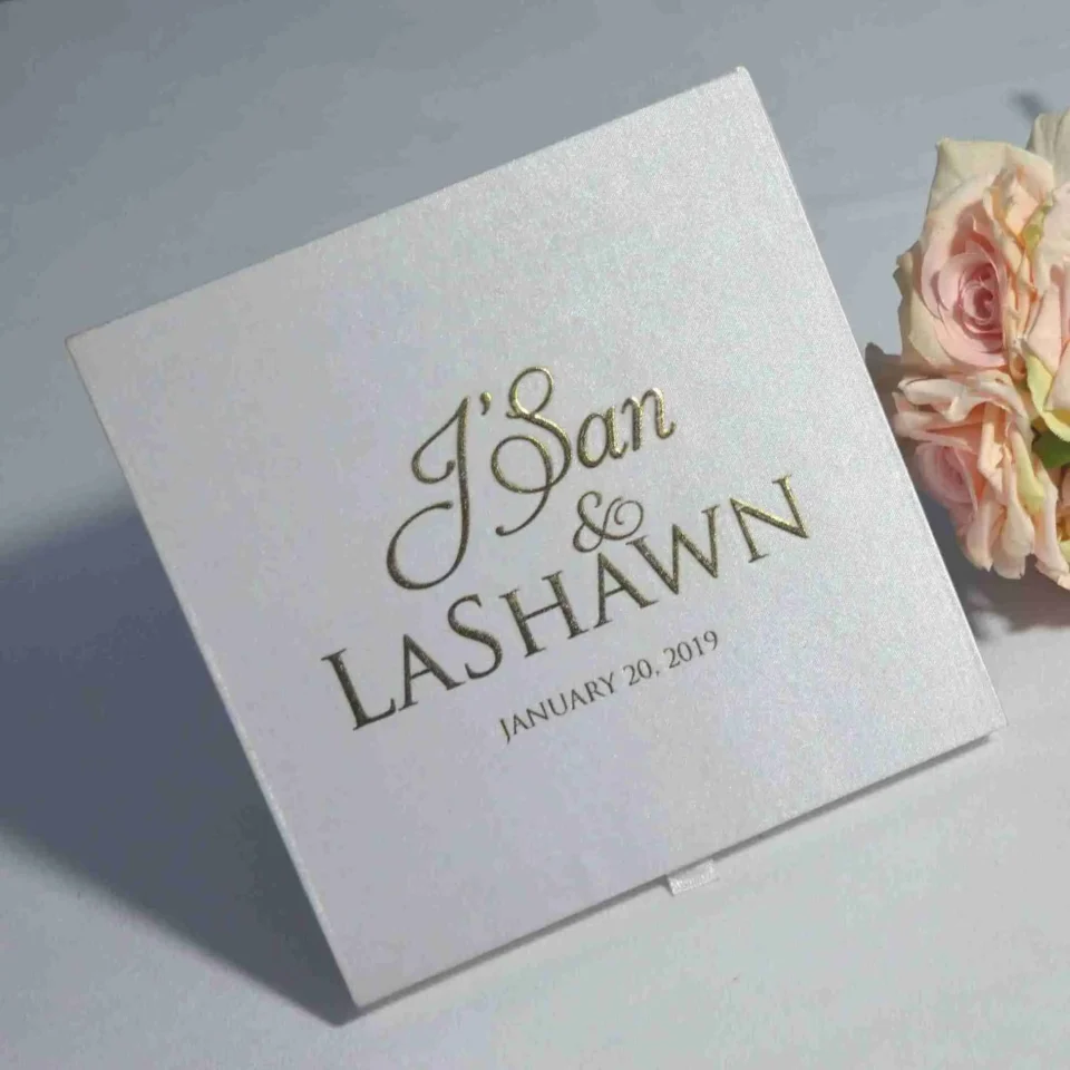 Satin wedding invitation box with gold foil names and date, styled beside blush pink roses