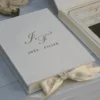 Close-up of a closed ivory velvet wedding invitation box with a gold foil monogram and names on the lid, with an open acrylic invitation box behind it.