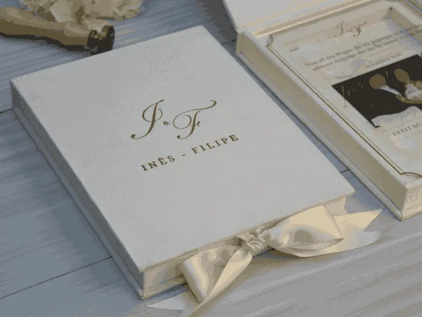 Close-up of a closed ivory velvet wedding invitation box with a gold foil monogram and names on the lid, with an open acrylic invitation box behind it.