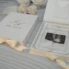 Closed ivory velvet wedding invitation box with a gold foil monogram beside an open box showing a clear acrylic invitation layered over a couple’s photo, with cream flowers.