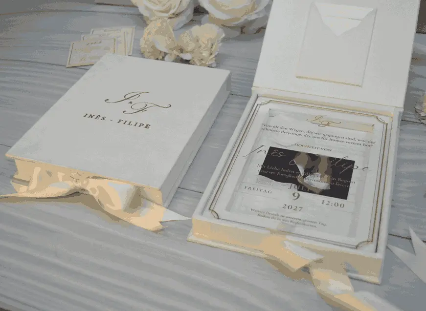 Closed ivory velvet wedding invitation box with a gold foil monogram beside an open box showing a clear acrylic invitation layered over a couple’s photo, with cream flowers.