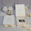 Closed ivory velvet wedding invitation box with a gold monogram beside an open box with an RSVP card in the lid pocket and a clear acrylic invitation, with white roses.