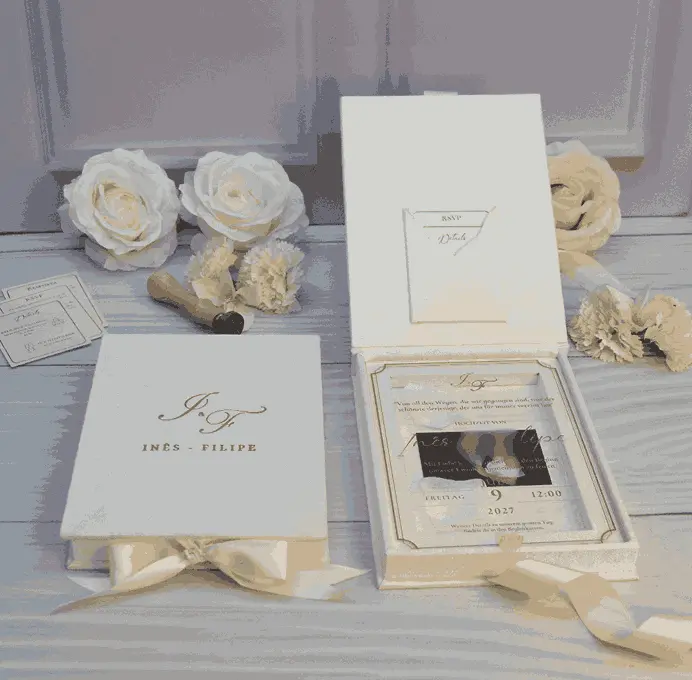 Closed ivory velvet wedding invitation box with a gold monogram beside an open box with an RSVP card in the lid pocket and a clear acrylic invitation, with white roses.